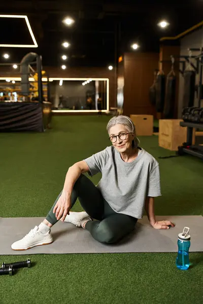 stock image joyous pretty mature woman in sportswear and glasses sitting on floor and smiling happily at camera