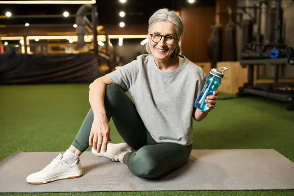 stock image good looking jolly mature sportswoman with glasses holding water bottle and smiling at camera
