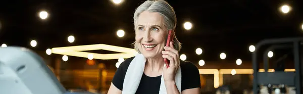 stock image positive senior woman with towel on shoulders talking by phone while on treadmill in gym, banner