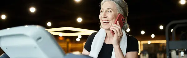 stock image jolly mature woman with towel on shoulders talking by phone while on treadmill in gym, banner