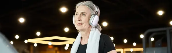 stock image mature positive woman with gray hair and headphones exercising on treadmill in gym, banner