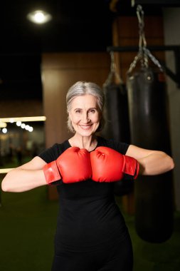 Spor kıyafetli, gri saçlı, spor salonunda boks eldivenleriyle poz veren olgun, neşeli bir kadın.