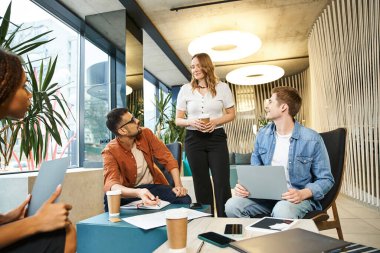 A diverse group of individuals sit around a table, laptops open, collaborating on projects in a coworking space. clipart