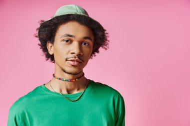 Kıvırcık saçlı, yeşil tişörtlü ve uyumlu şapkalı neşeli Afro-Amerikan bir adam canlı pembe bir arka planda.