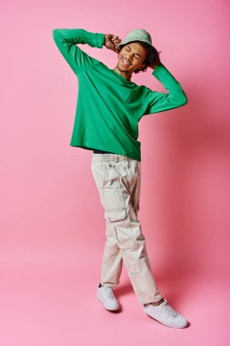 Kıvırcık saçlı, yeşil tişörtlü, beyaz pantolonlu, pembe arka planda coşkulu, neşeli Afro-Amerikan bir adam..