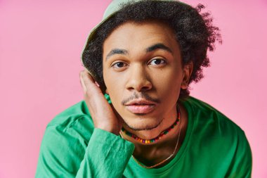 young African American man with curly hair wearing a green shirt and hat, on a pink background. clipart