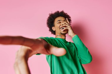 Yeşil tişörtlü, pembe arka planda gülümseyen neşeli, Afro-Amerikan bir adam..