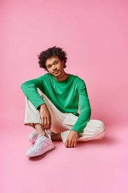 Yeşil gömlekli, beyaz pantolonlu, pembe arka planda kendine güveni olan neşeli, Afro-Amerikan bir adam..