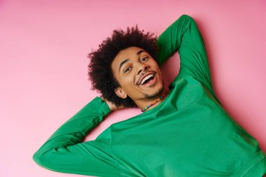 Neşeli, kıvırcık saçlı, pembe arka planda uzanan Afro-Amerikan adam, bir memnuniyet hissi sergiliyor..