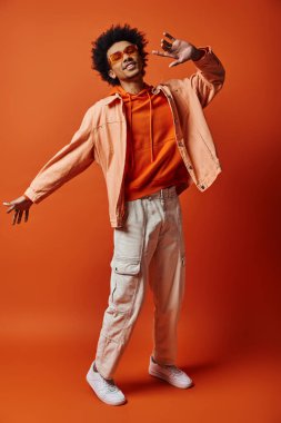 Kıvırcık saçlı, modaya uygun genç bir Afro-Amerikan erkek canlı turuncu bir arka plana karşı gömlek ve beyaz pantolon giyiyor..
