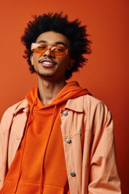 Ceket ve güneş gözlüğü takan modaya uyan Afrikalı Amerikalı bir adam turuncu arka planda tutum ve duygu yayıyor..