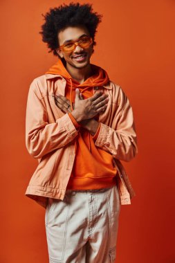 Kıvırcık saçlı, turuncu ceketli ve güneş gözlüklü genç bir Afrikalı Amerikalı parlak turuncu arka planda duruyor..