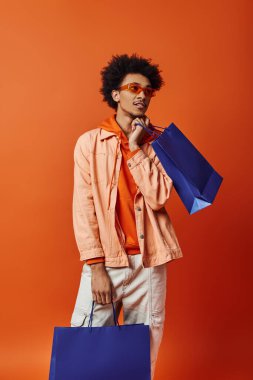 Mavi bir alışveriş çantası tutan ve turuncu arka plandaki kameraya bakan şık Afro-Amerikan bir adam..