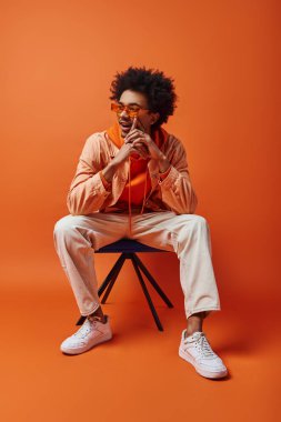 Kıvırcık saçlı ve güneş gözlüklü şık, genç bir Afro-Amerikalı, turuncu arka planda bir sandalyenin üzerinde cesurca oturuyor..