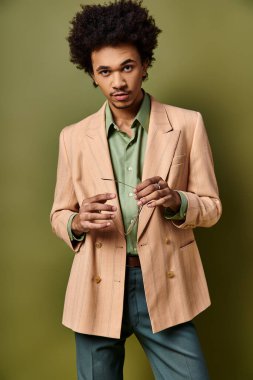 Şık, kıvırcık saçlı, kahverengi ceketli ve yeşil gömlekli, modaya uygun giyinmiş ve güneş gözlüklü Afro-Amerikan bir adam..