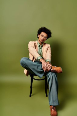 Şık, kıvırcık saçlı, güneş gözlüklü, ayakları çapraz bir sandalyede oturan Afro-Amerikan bir adam..