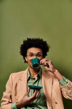 Moda giysileri ve güneş gözlüğü takan, yeşil arka planda bir fincan kahve içen şık, genç bir Afrikalı Amerikalı..