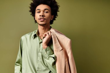 Stylish young African American man with curly hair holds a jacket over his shoulder, wearing trendy attire on a green background. clipart