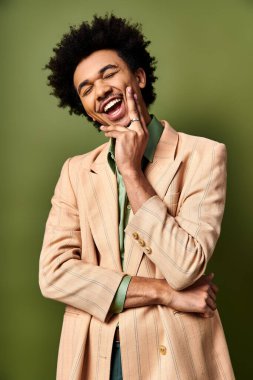 Young African American man in stylish suit laughs joyfully, holding hand to face against vibrant green backdrop. clipart