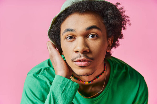 young African American man with curly hair wearing a green shirt and hat, on a pink background.