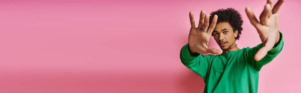 Cheerful curly African American man in casual green shirt with hands raised, expressing positivity on a pink background.