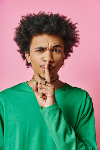 Curly African American man in casual wear looks pensive, with finger in mouth, against pink background.