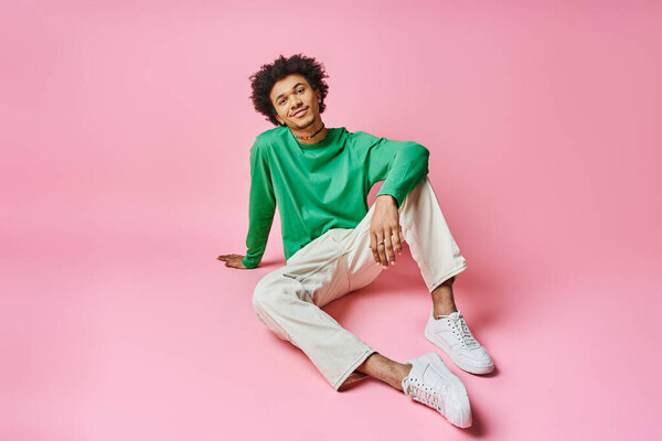 Веселый афроамериканец с вьющимися волосами, сидящий на розовой поверхности, одетый в зеленую рубашку и белые штаны.