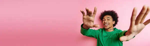stock image A cheerful young African American man with curly hair in casual wear raising his hands in jubilation on a pink background.
