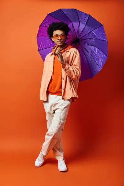 stock image A young African American man with curly hair holds a purple umbrella against a bold orange backdrop, exuding style and personality.