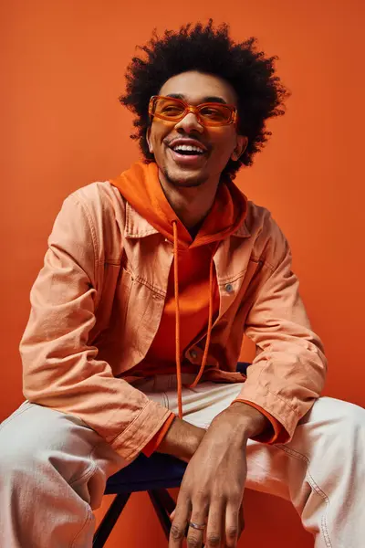 stock image Young African American man with curly hair sitting confidently on chair, sporting stylish sunglasses against orange background.