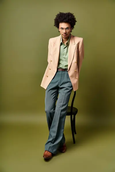 stock image A stylish young African American man in a suit and sunglasses stands confidently next to a chair against a green background.