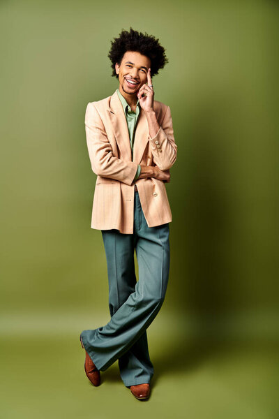 A trendy young African American man with curly hair wearing a tan jacket, green pants against a green backdrop.