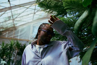 A stylish African American man stands in a greenhouse, holding his hand to his head. clipart