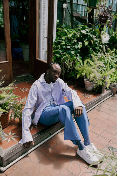 Красивый афроамериканец сидит на скамейке перед яркими зелеными растениями.
