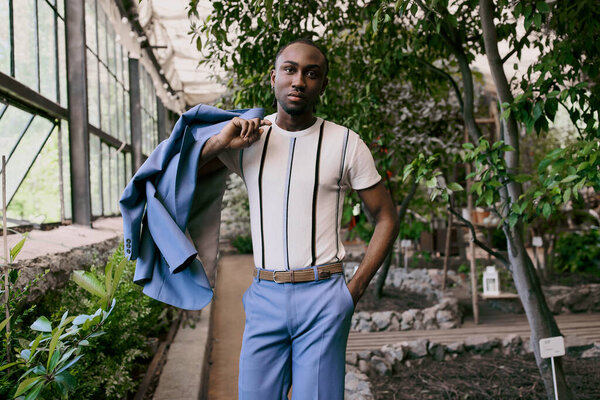 Афроамериканец в белой рубашке и синих штанах позирует в зеленом саду.