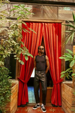 A dapper African American man strikes a pose in front of a vibrant red curtain. clipart