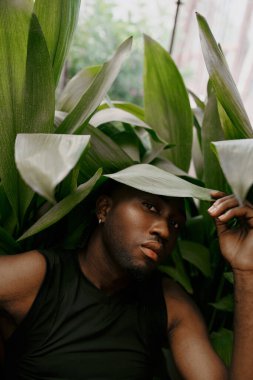 Yakışıklı Afrikalı Amerikalı adam yeşil bir bitkinin önünde şık bir şekilde oturuyor..