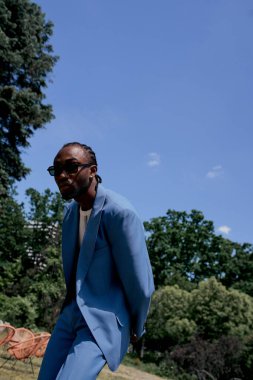 Mavi takım elbiseli ve güneş gözlüklü şık Afro-Amerikan adam yemyeşil bir bahçede poz veriyor..