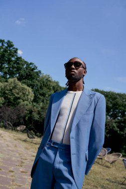 Mavi takım elbiseli ve güneş gözlüklü Afro-Amerikan bir adam yemyeşil bahçede poz veriyor..