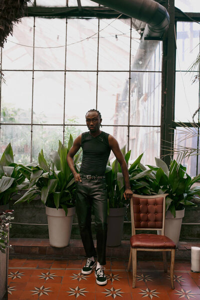 Стильный мужчина, стоящий у окна со стулом поблизости.