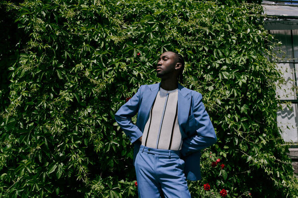 An African American man in a blue suit stands confidently in front of a lush green bush.