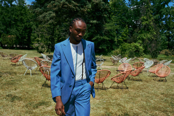 An African American man in a stylish blue suit strolls through a lush green field.