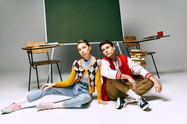 İki öğrenci üniversite sınıfında yeşil bir panonun önünde günlük kıyafetleriyle oturuyorlar..