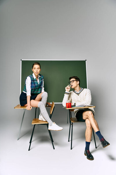 A stylish man and woman sit in front of a green board in a college setting.