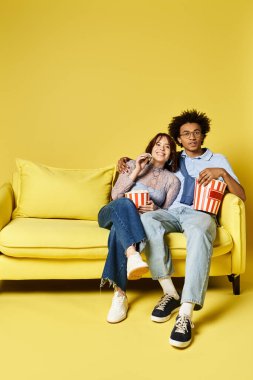 Şık giyinmiş bir adam ve bir kadın kanepede oturup sıcak bir ortamda patlamış mısırın tadını çıkarıyorlar..
