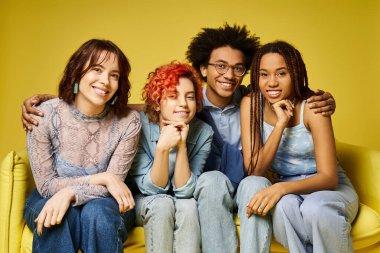 Young multicultural friends in stylish attire relax on a yellow couch in a studio setting. clipart