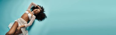 Modayı takip eden mayo giymiş Afro-Amerikalı kadın mavi arka planda rahatlıyor.