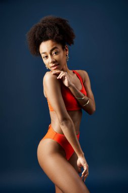 Göz kamaştırıcı, turuncu bikinili Afro-Amerikalı kadın canlı mavi bir arka planda poz veriyor..