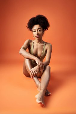 Afro-Amerikan kadın şık bikinili, canlı turuncu arka planda poz veriyor..