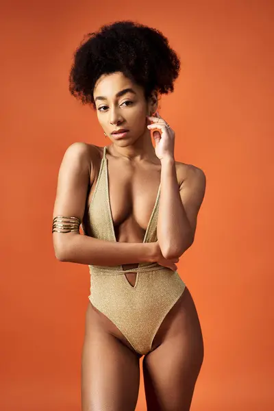 Stylish African American woman in gold swimsuit striking a pose on vibrant orange background.